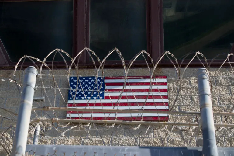 An American flag is seen through razor wire on the control tower of the Camp VI detention facility at the Guantanamo Bay Naval Base, Cuba, on April 17, 2019 [Alex Brandon/AP Photo]