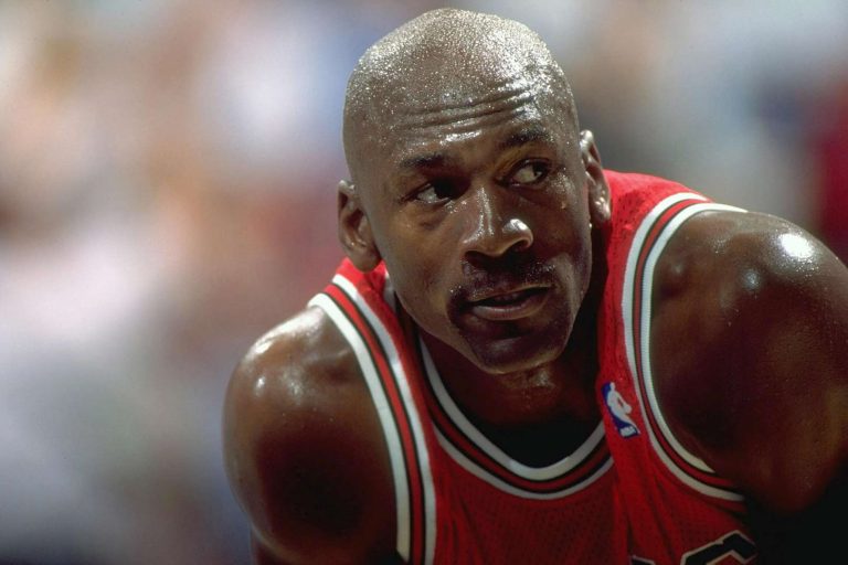 A close-up photograph of a basketball player in a red Chicago Bulls jersey, wearing a look of intense concentration during a game, with sweat visible on his face and bald head. His red uniform features the iconic Bulls striping around the neckline.